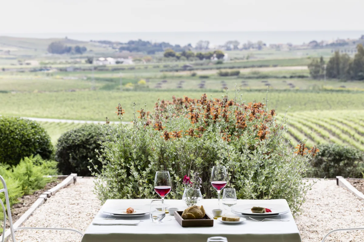 Table with wine glasses set in the garden of La Foresteria Planeta Estate, with vineyards beyond