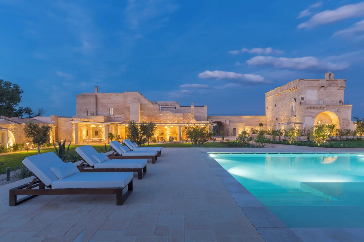 Masseria Donna Menga at dusk, its lit stone walls beyond a row of loungers by the pool