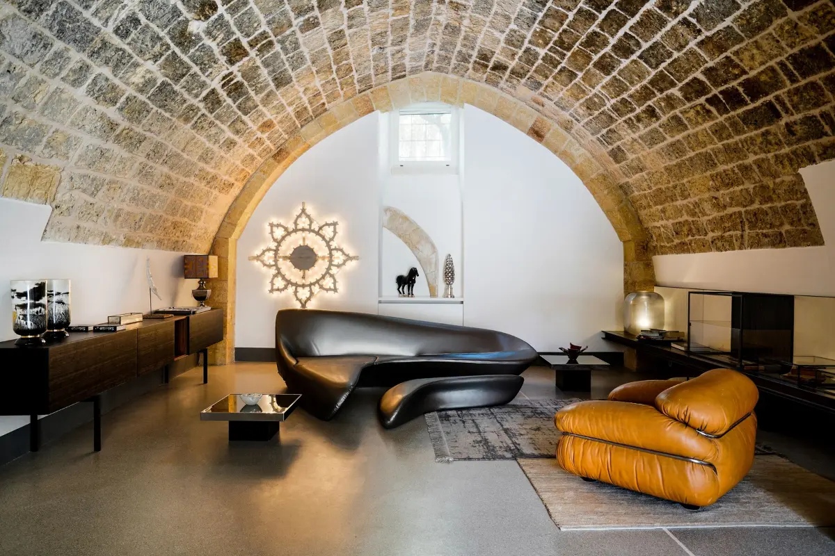 Suite at Palazzo De Noha in Lecce under a vaulted stone ceiling, with a black sculpted sofa, a tan leather armchair and a sunburst mirror